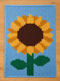 A knitted rectangular chunky blanket featuring a large sunflower design. The sunflower has a brown center, yellow petals, and a green stem and leaves, set against a light blue background.
