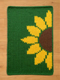A knitted green rectangular chunky blanket with a large sunflower design. The sunflower has yellow petals and a brown center. The blanket is made of thick yarn and has a textured appearance.
