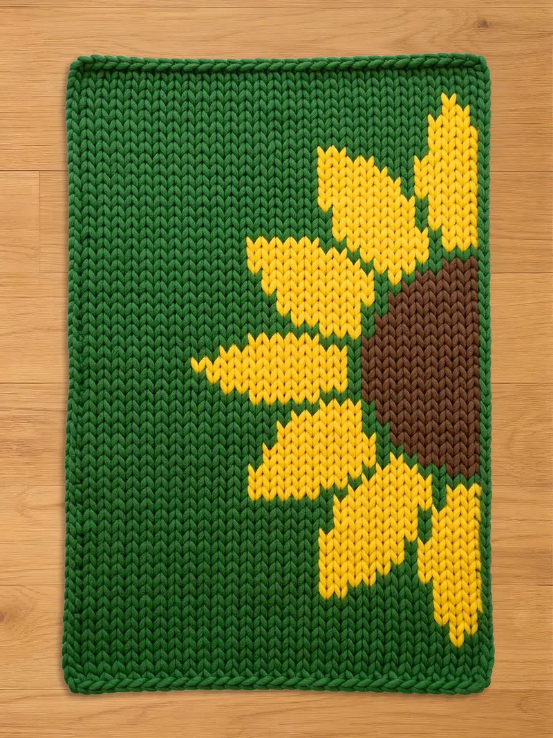 A knitted green rectangular chunky blanket with a large sunflower design. The sunflower has yellow petals and a brown center. The blanket is made of thick yarn and has a textured appearance.