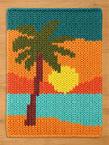 A rectangular knit chunky blanket featuring a colorful landscape design. The pattern displays a brown palm tree trunk with dark green fronds against a sunset background of yellow, orange, and light blue horizontal stripes.
