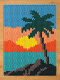 A rectangular chunky knit textile featuring a graphic design of a palm tree on a beach at sunset. The scene includes a blue sky, an orange horizon, a bright yellow sun, a teal ocean, and a dark gray sandy shore with a palm tree silhouette.