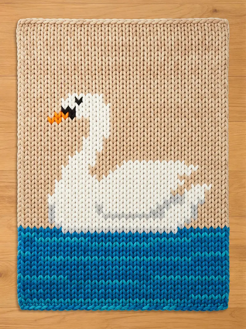 Knitted swan design on a rectangular chunky blanket. The swan is white with black and orange details, set against a beige background. The bottom features blue water. The blanket is made of thick, textured yarn.