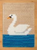 Knitted swan design on a rectangular chunky blanket. The swan is white with black and orange details, set against a beige background. The bottom features blue water. The blanket is made of thick, textured yarn.