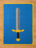Chunky knit blanket in bright blue featuring a large sword design with a silver blade, gold hilt, and brown handle, finished with a yellow border