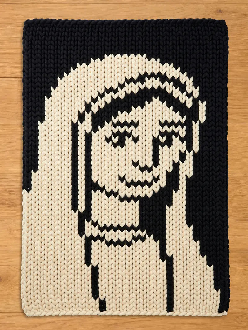 A rectangular, knitted chunky blanket featuring a pixelated image of a face. The face is rendered in black and cream, with a simple, geometric design. The blanket is made of thick yarn, giving it a textured appearance.