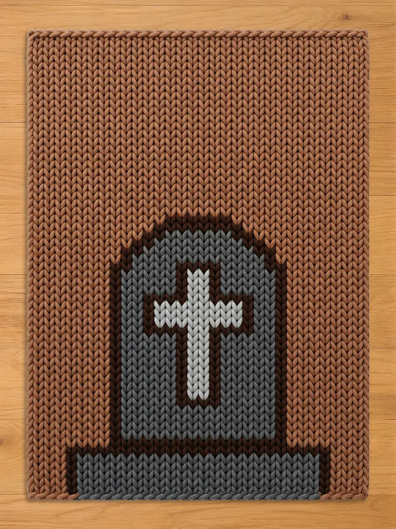A rectangular knitted chunky blanket featuring a brown background with a centered gray tombstone motif. The tombstone is outlined in dark brown and contains a white cross at its center, with a gray base at the bottom of the design.