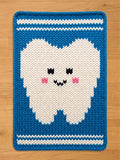 A rectangular, knitted chunky blanket featuring a smiling tooth design. The tooth is white with pink cheeks and black eyes and a mouth. The blanket has a blue border with white stripes at the top and bottom. The blanket is made of a soft, textured material.