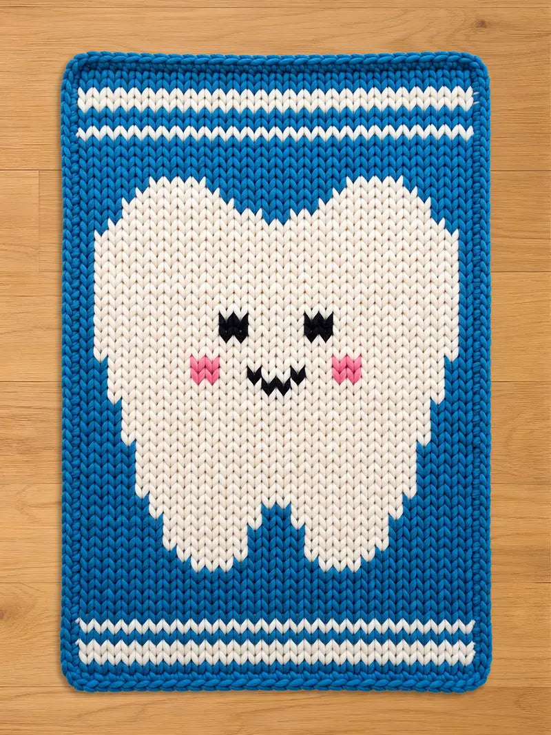 A rectangular, knitted chunky blanket featuring a smiling tooth design. The tooth is white with pink cheeks and black eyes and a mouth. The blanket has a blue border with white stripes at the top and bottom. The blanket is made of a soft, textured material.