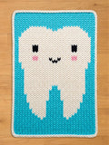 A knitted chunky blanket featuring a smiling tooth design. The tooth is white with black eyes, pink cheeks, and a black mouth, set against a turquoise background with a white border. The blanket is rectangular.