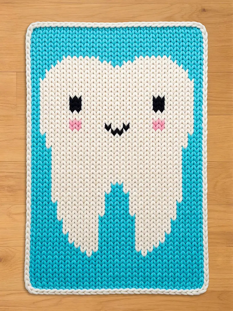 A knitted chunky blanket featuring a smiling tooth design. The tooth is white with black eyes, pink cheeks, and a black mouth, set against a turquoise background with a white border. The blanket is rectangular.