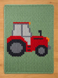 A knitted green blanket featuring a red tractor design. The tractor has gray and black wheels, a white and gray cab, and orange headlights. The blanket is made of a chunky knit material.