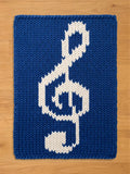 A chunky knit blanket in royal blue featuring a large white treble clef design in the center, laid flat on a light wooden surface.