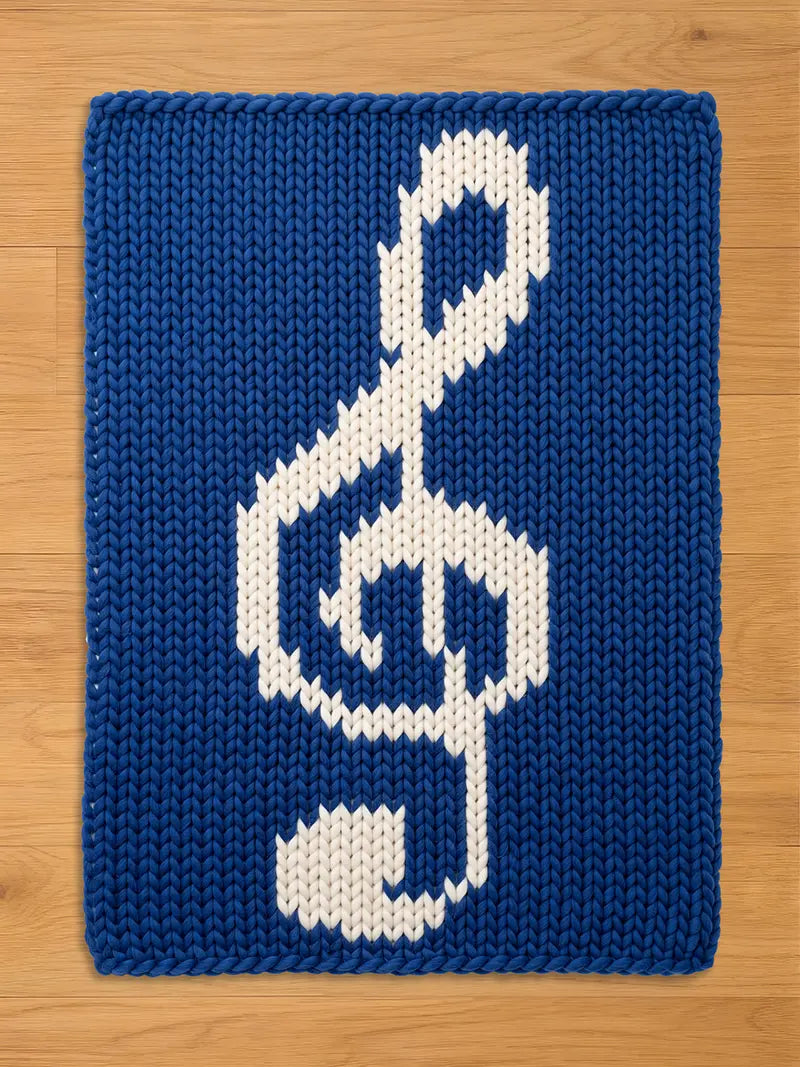 A chunky knit blanket in royal blue featuring a large white treble clef design in the center, laid flat on a light wooden surface.