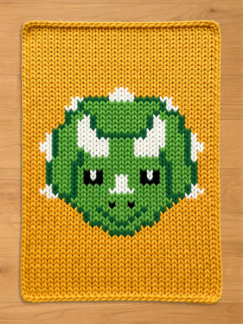 Mustard yellow chunky knit blanket featuring a large, central green triceratops dinosaur head design with white horns and black eyes