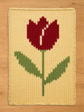 A knitted rectangular chunky blanket featuring a large tulip design. The tulip has a red flower with a white center, green stem and leaves, set against a yellow background. The blanket is made of a soft, textured material.