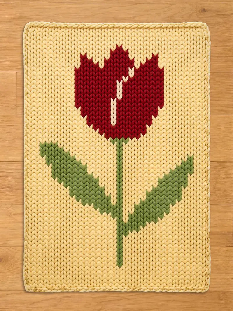 A knitted rectangular chunky blanket featuring a large tulip design. The tulip has a red flower with a white center, green stem and leaves, set against a yellow background. The blanket is made of a soft, textured material.