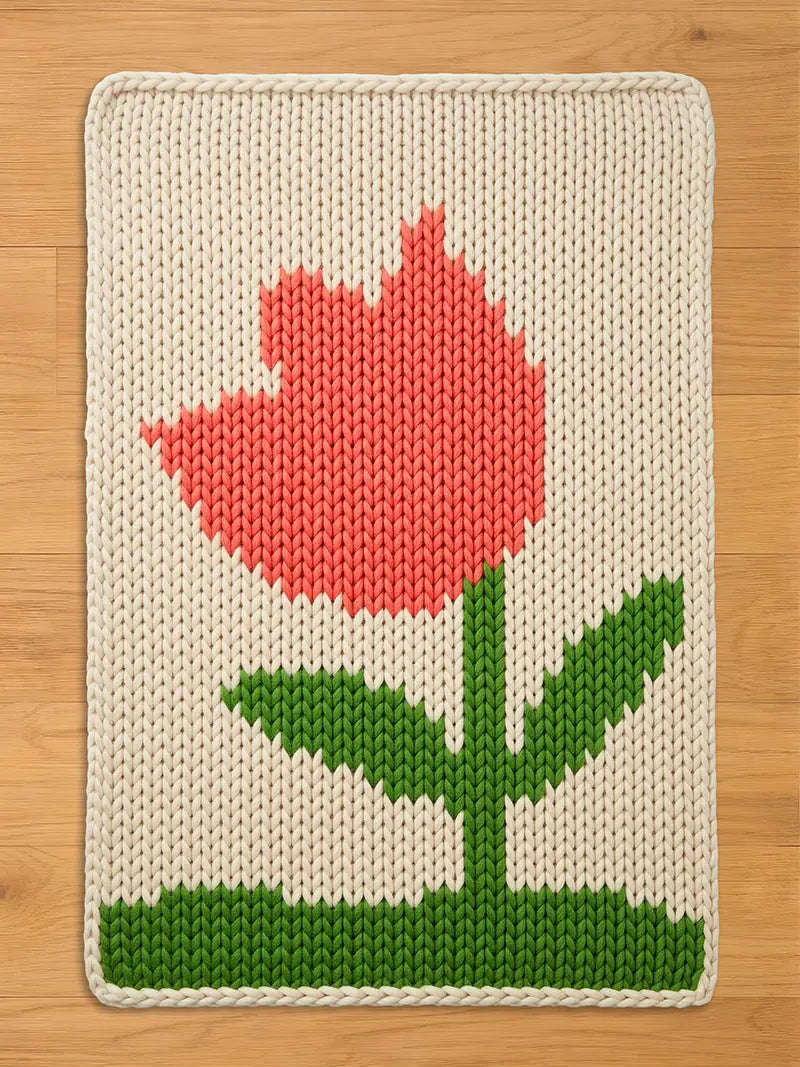 A knitted rectangular chunky blanket featuring a large, stylized tulip design. The flower is coral pink, with a green stem and leaves, set against an off-white background. The blanket has a textured, handmade appearance.
