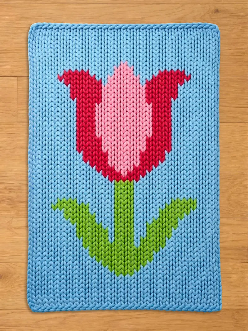 A knitted blue rectangular chunky blanket featuring a large tulip design. The tulip has a pink center and red petals, with a green stem and leaves. The blanket is made of a soft, textured material.