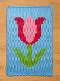A knitted blue rectangular chunky blanket featuring a large tulip design. The tulip has a pink center and red petals, with a green stem and leaves. The blanket is made of a soft, textured material.