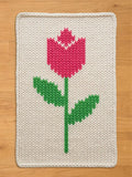 A knitted rectangular chunky blanket featuring a pink tulip with green leaves and stem on a cream background. The edges are neatly finished with a cream border, creating a cozy, handmade aesthetic.