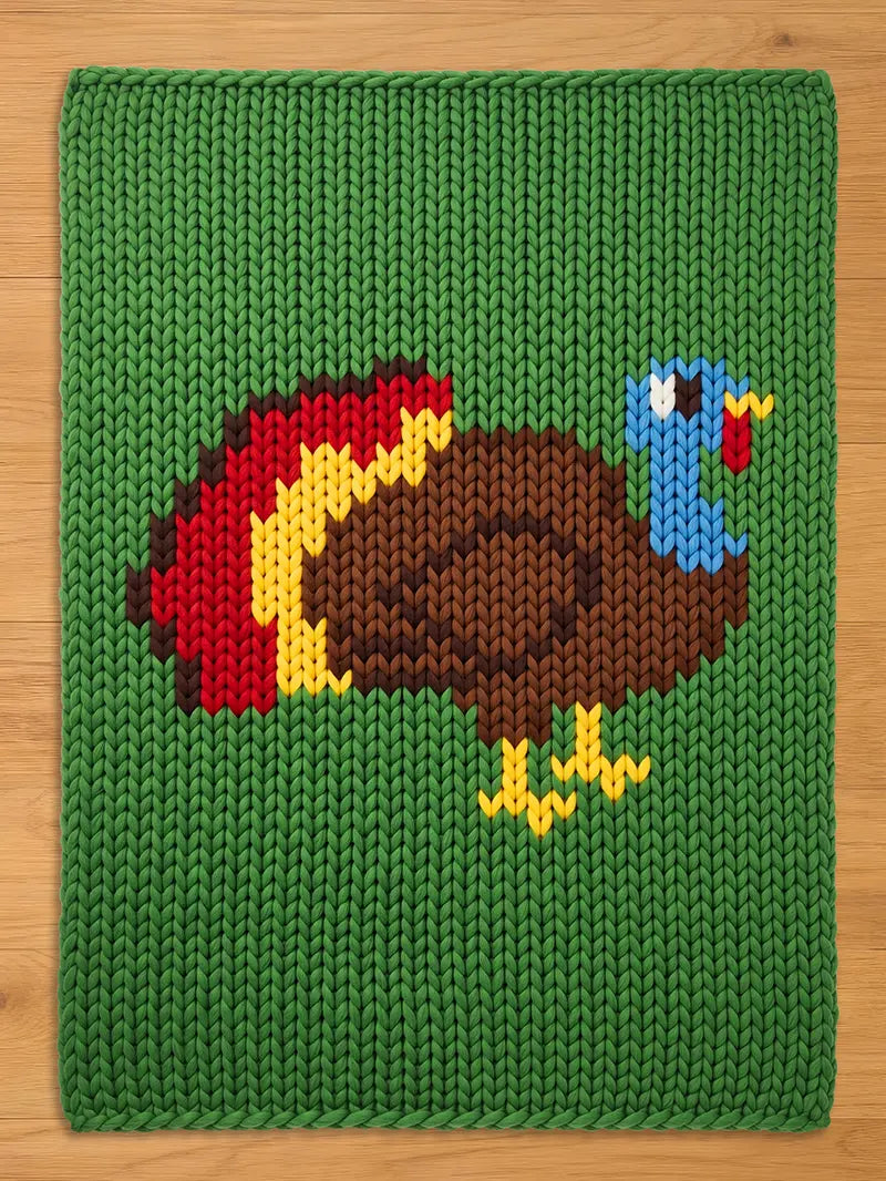 A knitted green chunky blanket featuring a colorful turkey design. The turkey has a brown body, blue head, and a red wattle. Its tail feathers are red, yellow, and brown. The blanket is made of a thick, textured yarn.