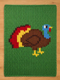 A knitted green chunky blanket featuring a colorful turkey design. The turkey has a brown body, blue head, and a red wattle. Its tail feathers are red, yellow, and brown. The blanket is made of a thick, textured yarn.