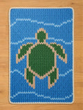 A knitted chunky blanket featuring a green turtle swimming in blue water. The turtle is outlined in a light tan color, and the water has white wave patterns. The blanket has a white border.
