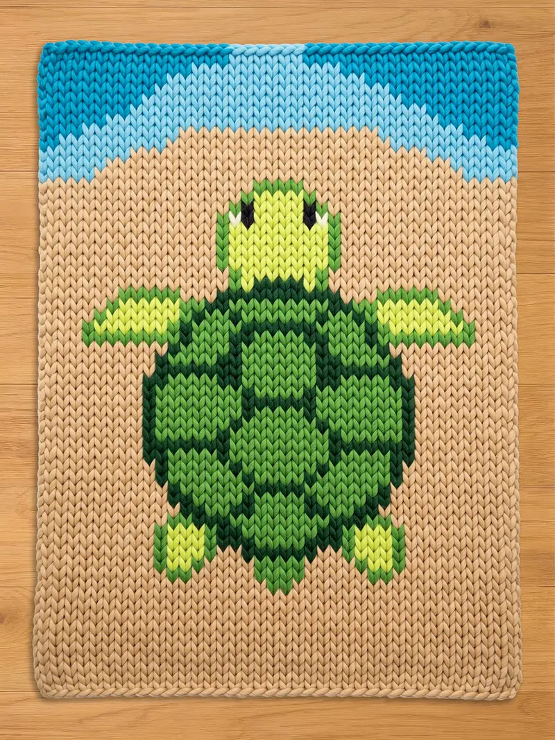 A rectangular chunky knit blanket featuring a central sea turtle graphic in shades of green and yellow. The turtle is set against a tan background with a blue arched border at the top, all created with a chunky, visible yarn stitch pattern.
