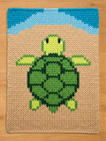 A rectangular chunky knit blanket featuring a central sea turtle graphic in shades of green and yellow. The turtle is set against a tan background with a blue arched border at the top, all created with a chunky, visible yarn stitch pattern.