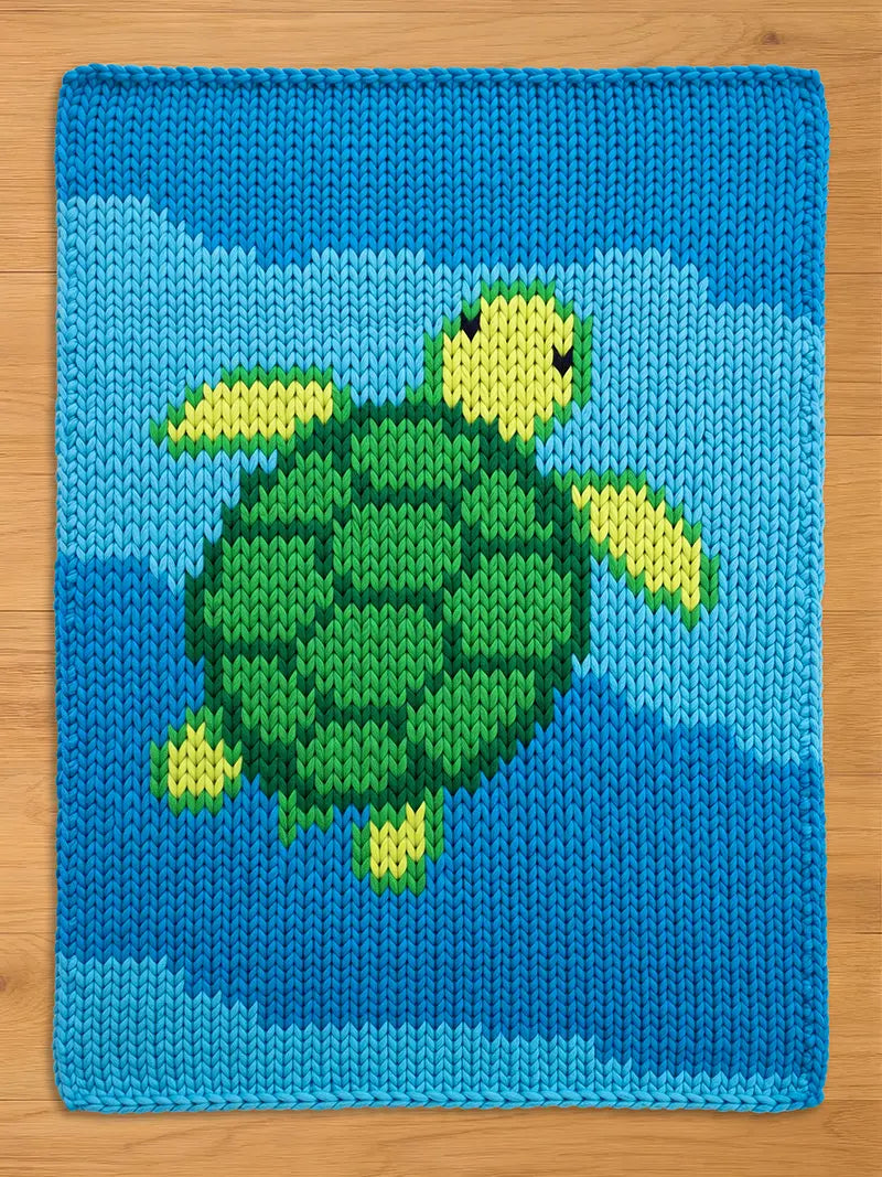 A knitted chunky blanket featuring a green and yellow turtle swimming in a blue ocean. The turtle has a textured shell and flippers. The background is a gradient of blue hues, creating a water effect.