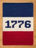 A rectangular, chunky knitted blanket in red, white, and blue. The top section is blue, the middle is white with the number "1776" in blue, and the bottom is red. The blanket is made of thick yarn.