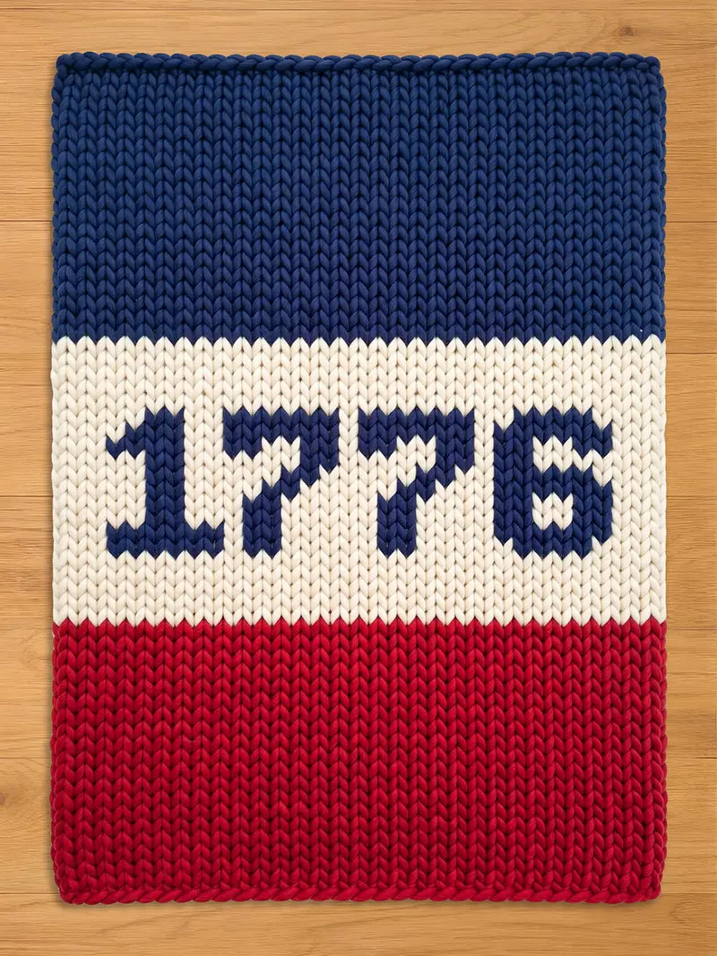 A rectangular, chunky knitted blanket in red, white, and blue. The top section is blue, the middle is white with the number "1776" in blue, and the bottom is red. The blanket is made of thick yarn.