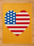 a rectangular chunky knit blanket laid flat on a wooden surface. The blanket features a solid mustard-yellow background with a large, central heart shape. The heart is patterned after the American flag, containing a blue upper-left section with white knit details representing stars, and red and white horizontal stripes filling the remainder of the heart.