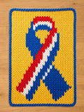 A knitted rectangular chunky blanket featuring a ribbon design in red, white, and blue against a yellow background. The blanket has a blue border and is made of thick yarn, suitable for home decor.