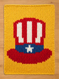 A rectangular chunky knit blanket in mustard yellow featuring a central graphic of a red, white, and blue top hat. The hat design includes vertical white stripes on a red background with a blue band and a single white star in the center.