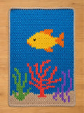 A knitted rectangular chunky blanket with a blue background depicting an underwater scene. A yellow fish with orange fins swims above coral and plants in shades of red, green, and purple. The blanket has a beige border.