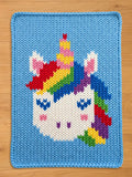 Light blue chunky knit blanket featuring a central white unicorn face design with closed blue eyes, rosy cheeks, and a vibrant rainbow mane