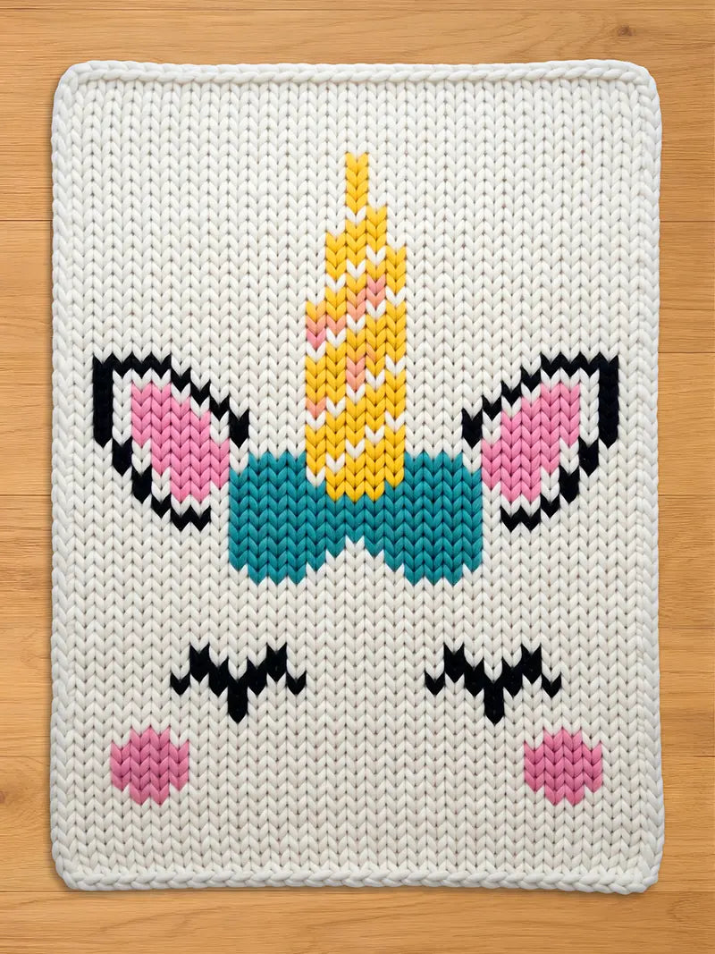 Chunky knit white blanket featuring a colorful unicorn face pattern with a yellow horn, pink ears, a teal bow, and sleepy black eyes