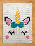 Chunky knit white blanket featuring a colorful unicorn face pattern with a yellow horn, pink ears, a teal bow, and sleepy black eyes