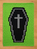 A rectangular knit blanket featuring a bright lime green background with a large, centered black coffin motif. The coffin is outlined in grey and contains a grey cross symbol in the center, all created with a chunky, visible knit texture.