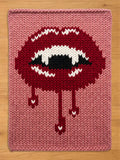 A rectangular chunky knit blanket featuring a large graphic of dark red lips with white fangs and black accents. The design includes red drips extending downward from the lips, all set against a solid dusty pink knitted background.