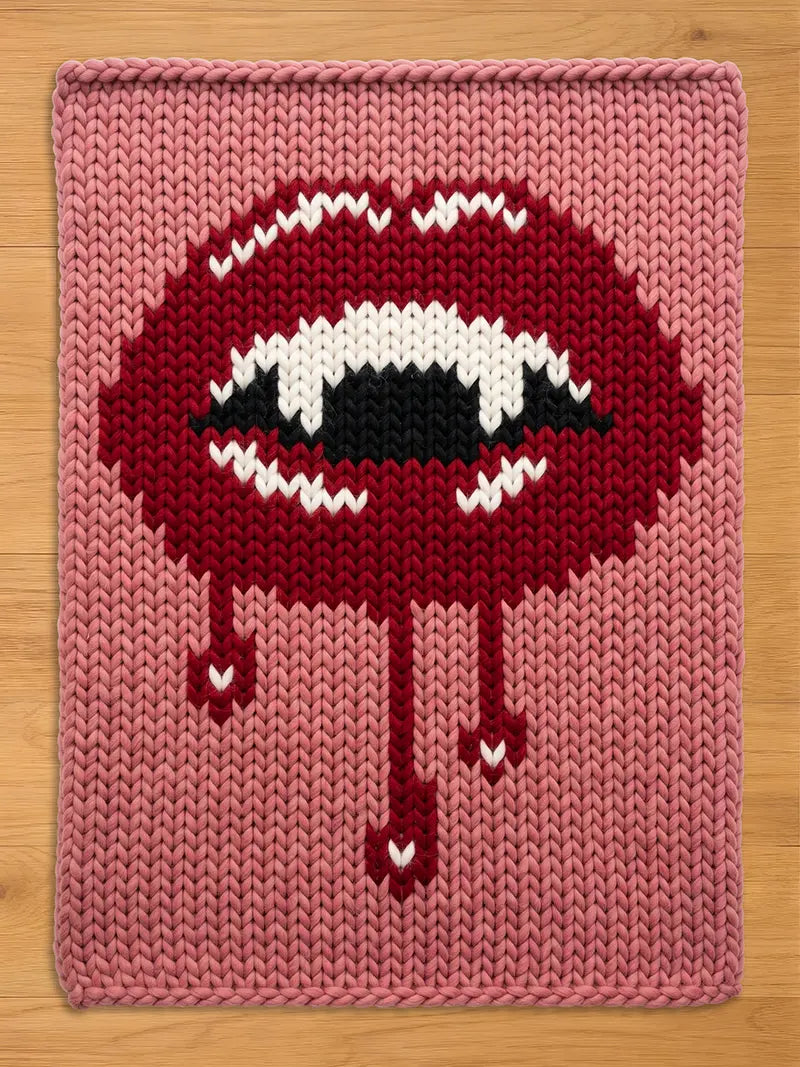 A rectangular chunky knit blanket featuring a large graphic of dark red lips with white fangs and black accents. The design includes red drips extending downward from the lips, all set against a solid dusty pink knitted background.