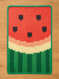 Rectangular chunky knit watermelon blanket laying flat on a wooden floor. The knitted pattern features a vibrant red center with black seed details, a curved pale yellow rind, and alternating dark and light green vertical stripes at the base