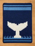 A dark blue chunky knit blanket featuring a large white whale tail silhouette in the center, bordered by a light blue wave pattern at the bottom and a solid light blue stripe at the top