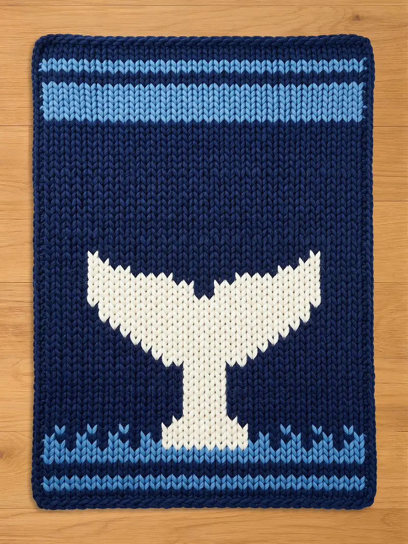 A dark blue chunky knit blanket featuring a large white whale tail silhouette in the center, bordered by a light blue wave pattern at the bottom and a solid light blue stripe at the top