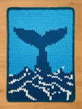A knitted chunky blanket with whale tail design in shades of blue and white. The whale tail is a darker blue against a light blue background. White accents create a wave pattern at the bottom. The edges are neatly finished with a light blue border.