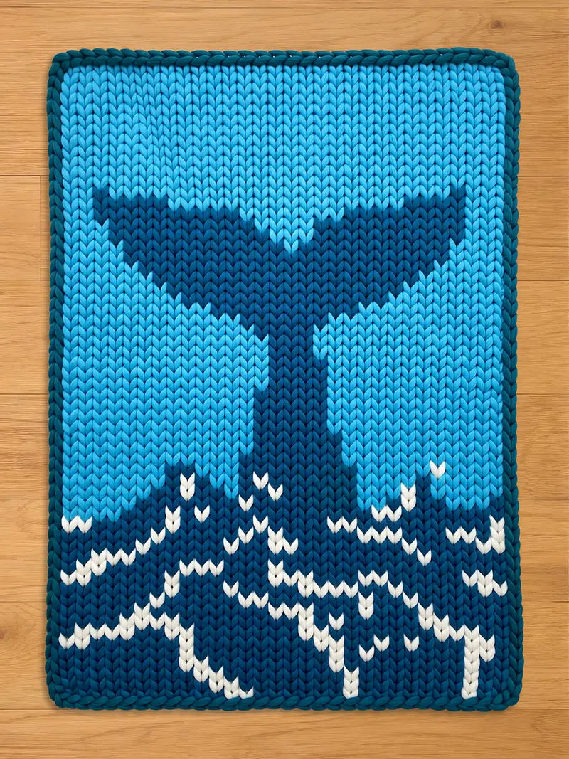 A knitted chunky blanket with whale tail design in shades of blue and white. The whale tail is a darker blue against a light blue background. White accents create a wave pattern at the bottom. The edges are neatly finished with a light blue border.