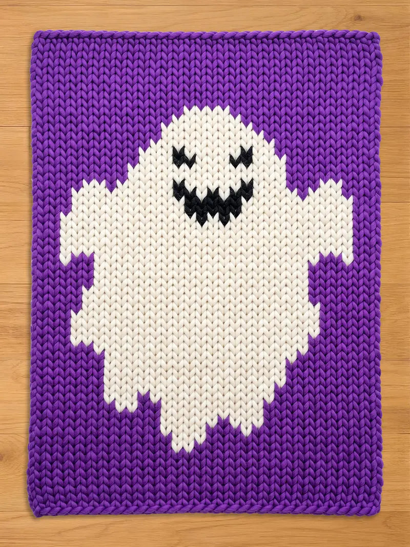  rectangular chunky knit blanket featuring a large white ghost motif centered on a solid purple background. The ghost has a jagged bottom edge and a black stitched face with two triangular eyes and a wide, toothy, smiling mouth.