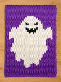  rectangular chunky knit blanket featuring a large white ghost motif centered on a solid purple background. The ghost has a jagged bottom edge and a black stitched face with two triangular eyes and a wide, toothy, smiling mouth.