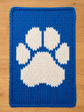 A blue knitted square chunky blanket with a large white paw print design. The blanket is made of thick yarn and has a textured appearance. The paw print is centered on the rug.